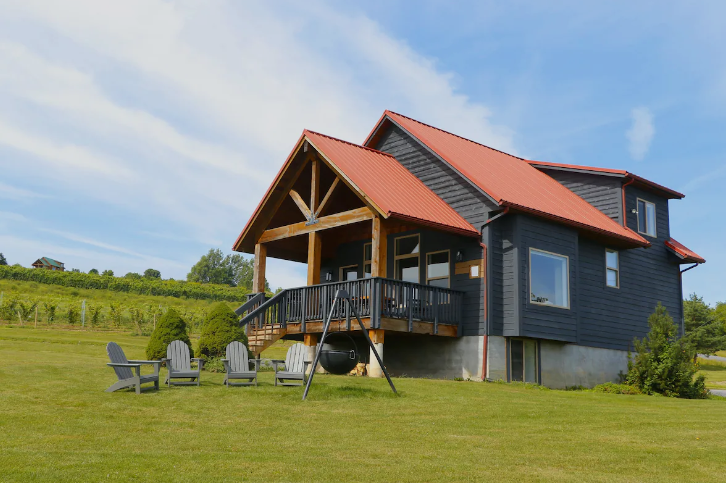 A dark gray house with a red roof, porch, swing, and Adirondack chairs on a grassy lawn.