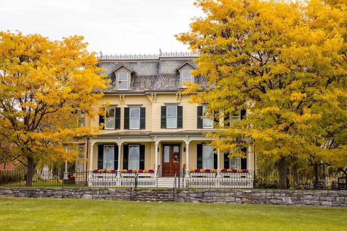 Victorian house with yellow autumn trees and a stone fence in front, on a grassy lawn.