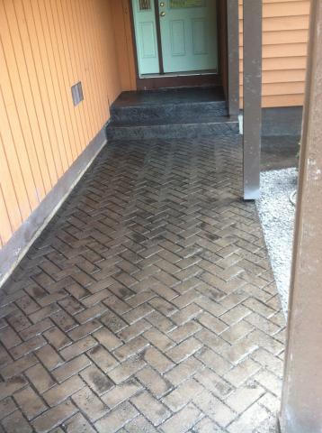 Brown herringbone brick walkway leading to green double doors on a house with orange siding.