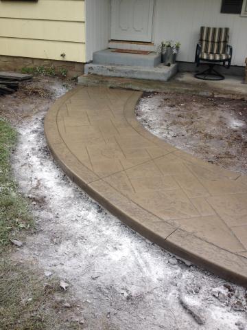 Curved, freshly-poured concrete walkway leading to a porch with a chair and potted plants.