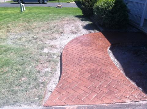 Curved brick walkway beside patchy grass and shrubs on a sunny day.