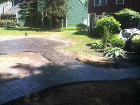 A newly paved stone walkway and patio in a sunny backyard with grass and garden plants.