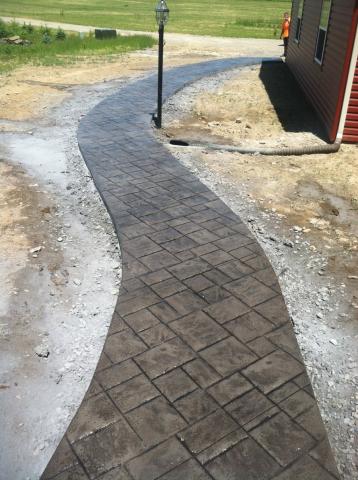 Curved, stamped concrete walkway with a lamp post beside a red building and grassy field.