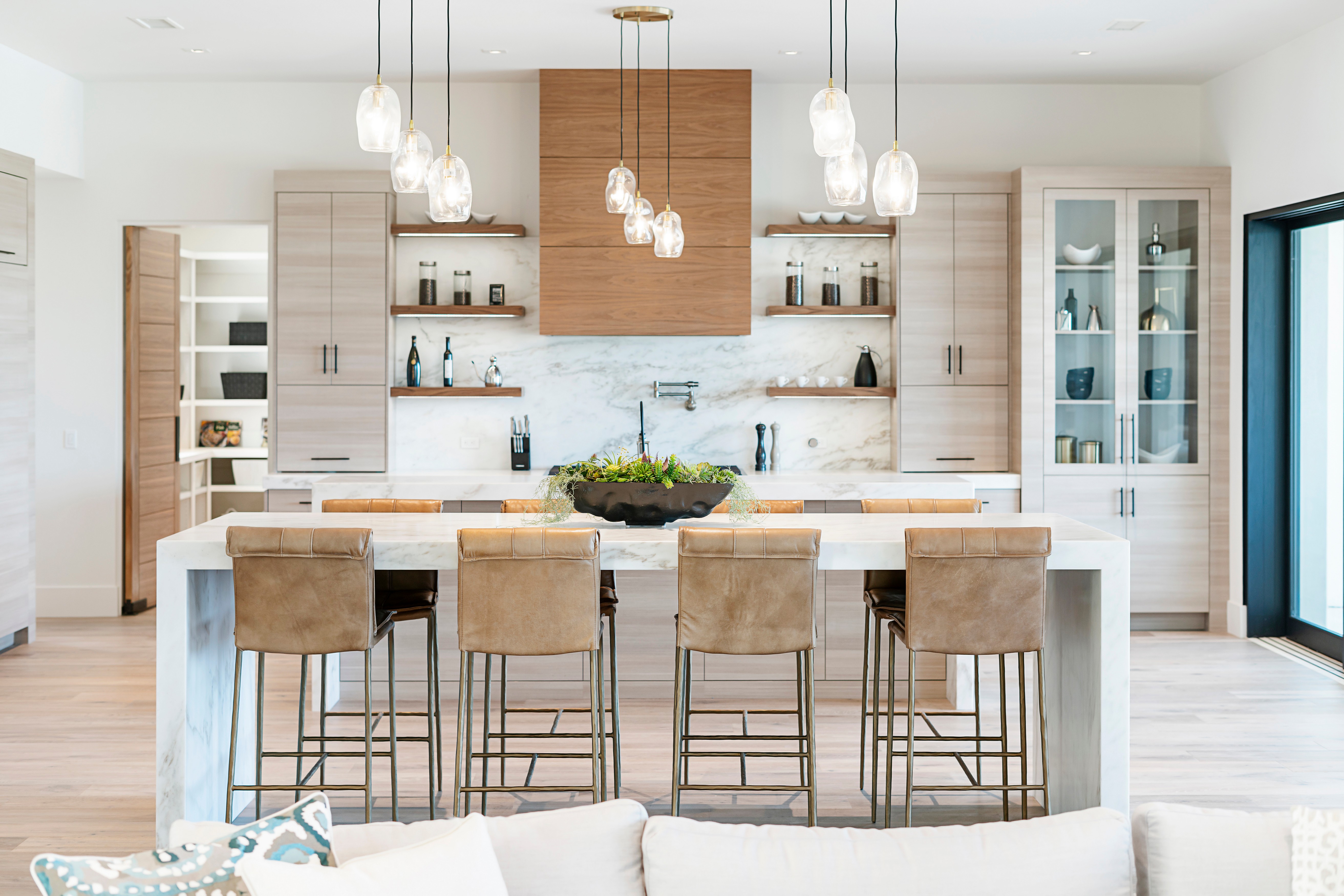 Modern kitchen with marble island, five brown barstools, pendant lights, and light wood cabinets.