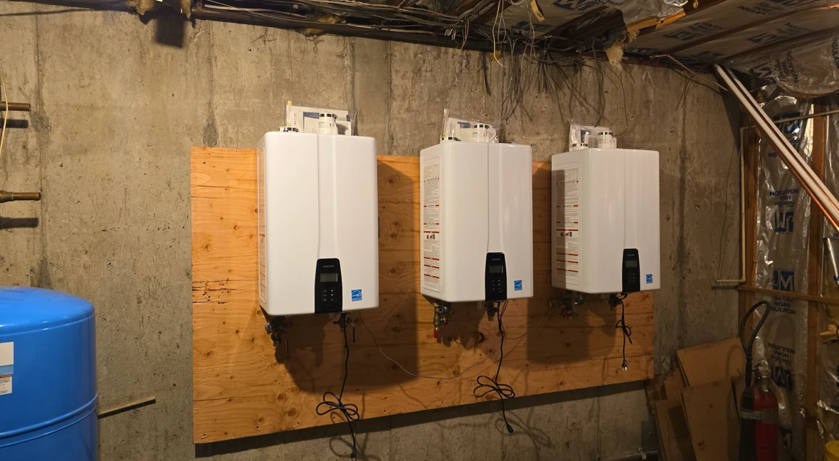 Three white tankless water heaters mounted on a plywood board in a basement utility room.