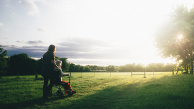 A younger woman pushes an elderly white-haired woman in a wheelchair into a grassy sunlit field that is surrounded by a small fence and a tall tree