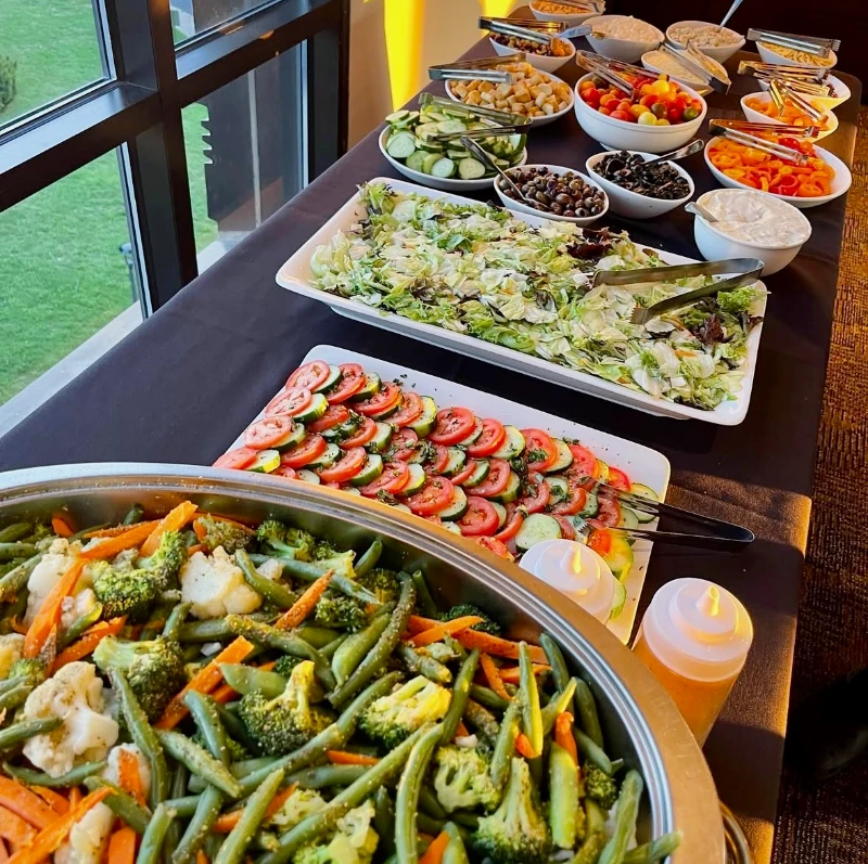 A buffet table with assorted salads, vegetables, and toppings next to a large window.