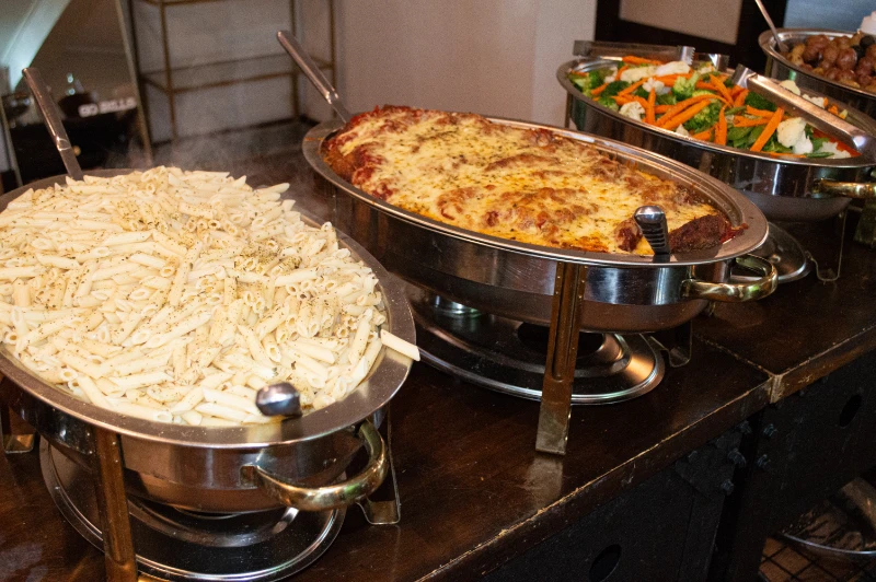 Buffet trays with penne pasta, baked lasagna, and mixed steamed vegetables on display.