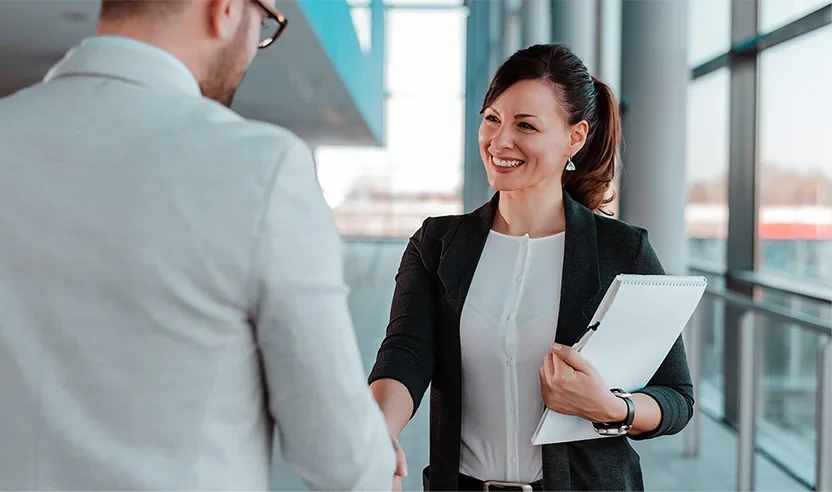 A woman holding documents smiles and shakes hands with a man in a bright office hallway.
