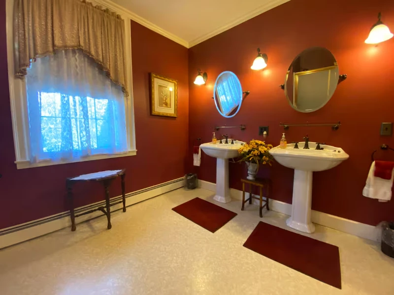 A spacious red bathroom with two pedestal sinks, oval mirrors, and a window with sheer curtains.