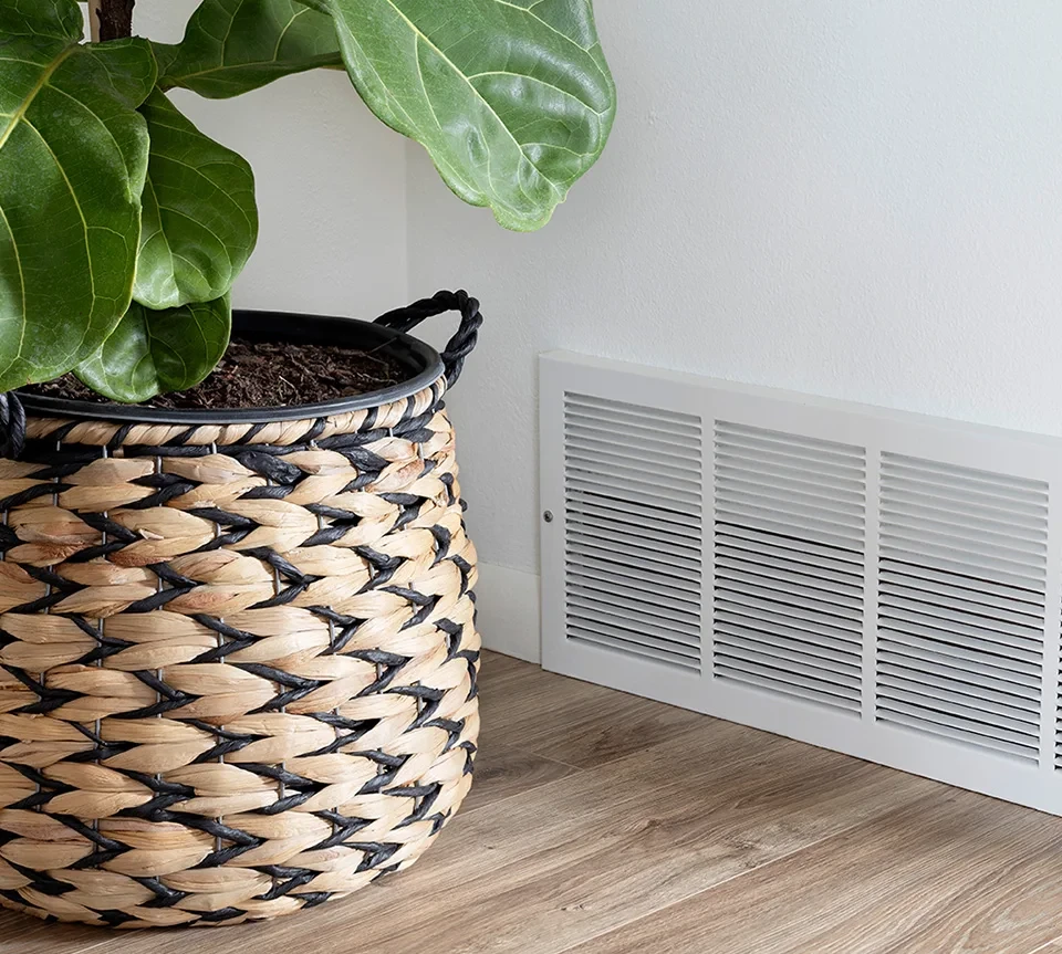 Basket with plant and air duct vent against a wall.