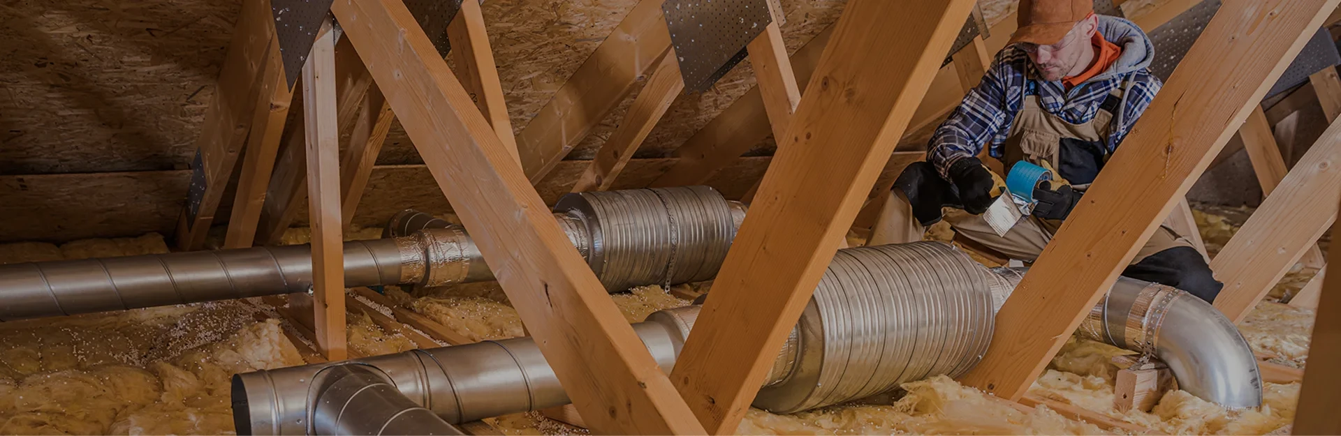 Worker in attic installing metal air ducts among wooden beams and insulation material.