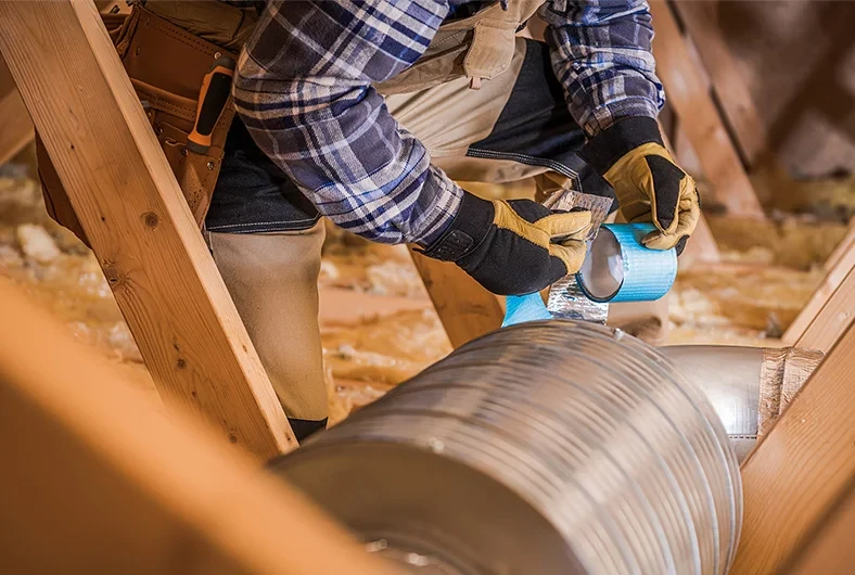 Person sealing an air duct in an attic with tape, wearing gloves and a plaid shirt.