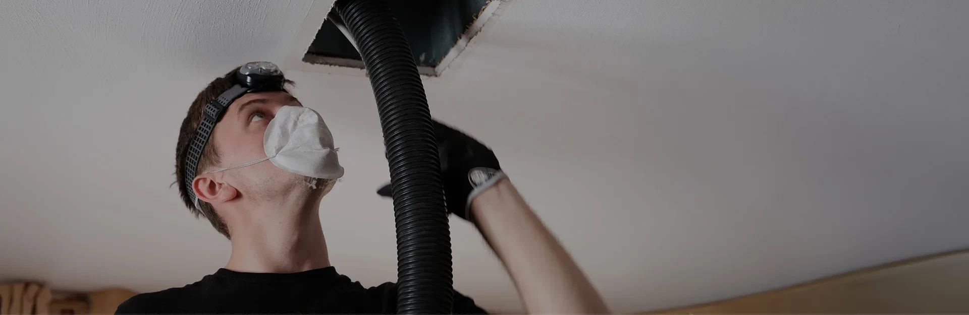A person in a mask and gloves cleans an air duct in the ceiling with a vacuum hose.