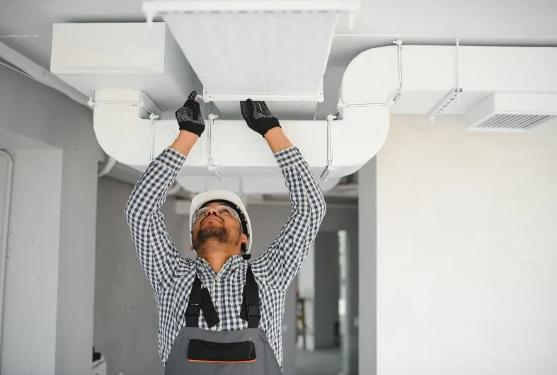 Worker in hard hat and gloves inspecting or installing ductwork on a ceiling indoors.