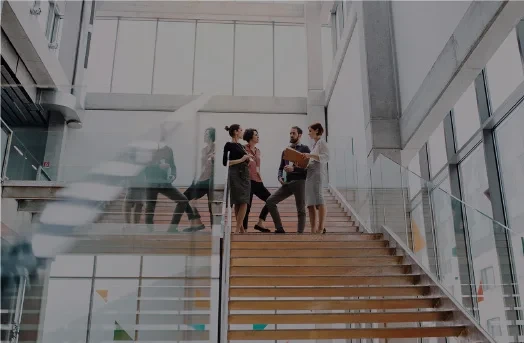 Five people stand and talk on a modern office staircase with glass railings and high windows.