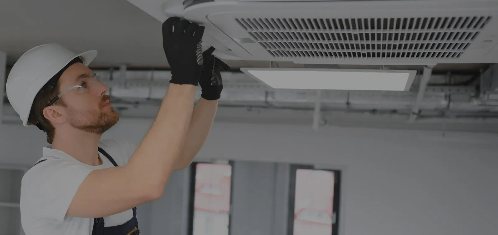 A worker in safety gear inspects or repairs a ceiling air conditioning vent indoors.