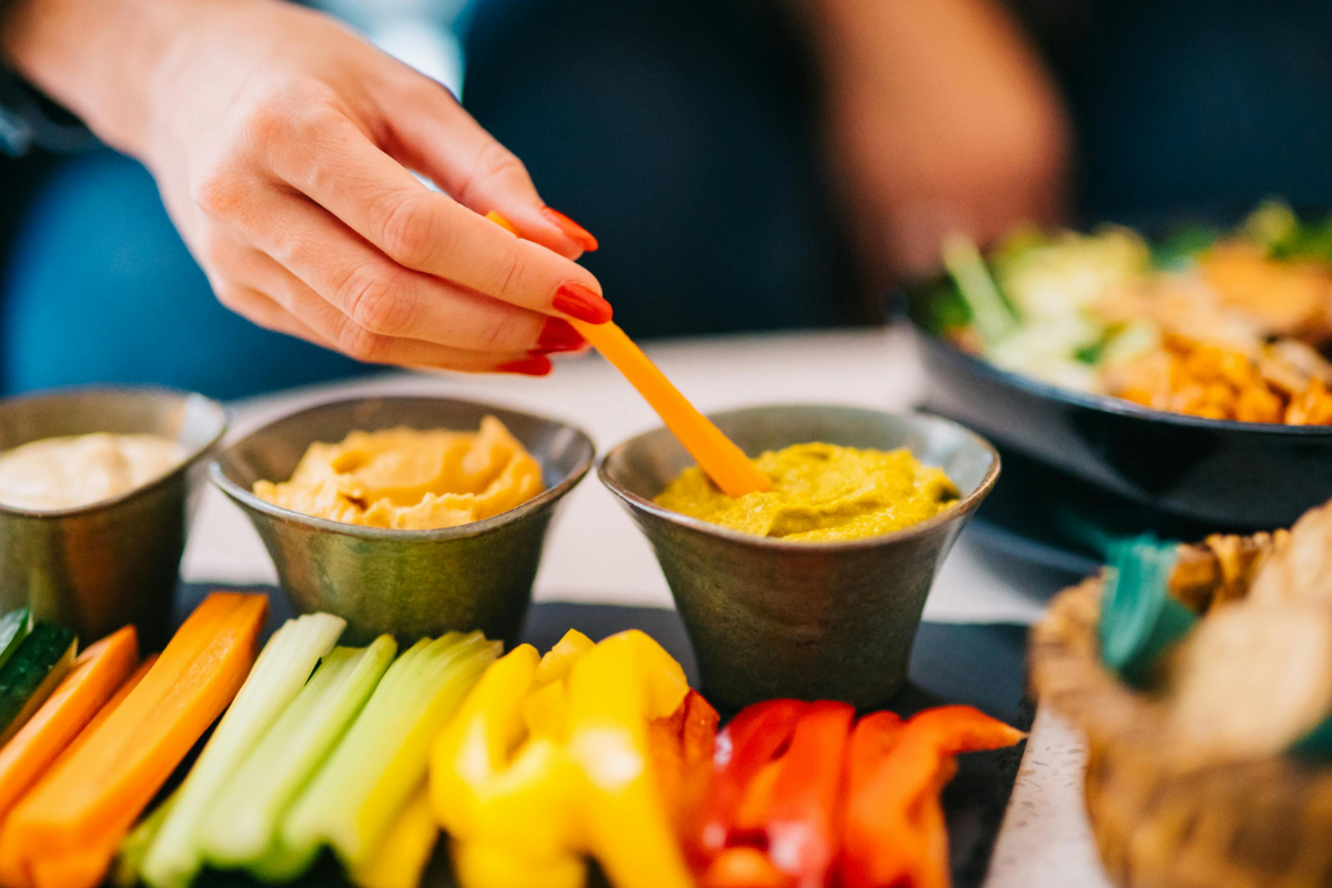 A hand dips a carrot stick into a bowl of yellow dip beside assorted sliced vegetables.
