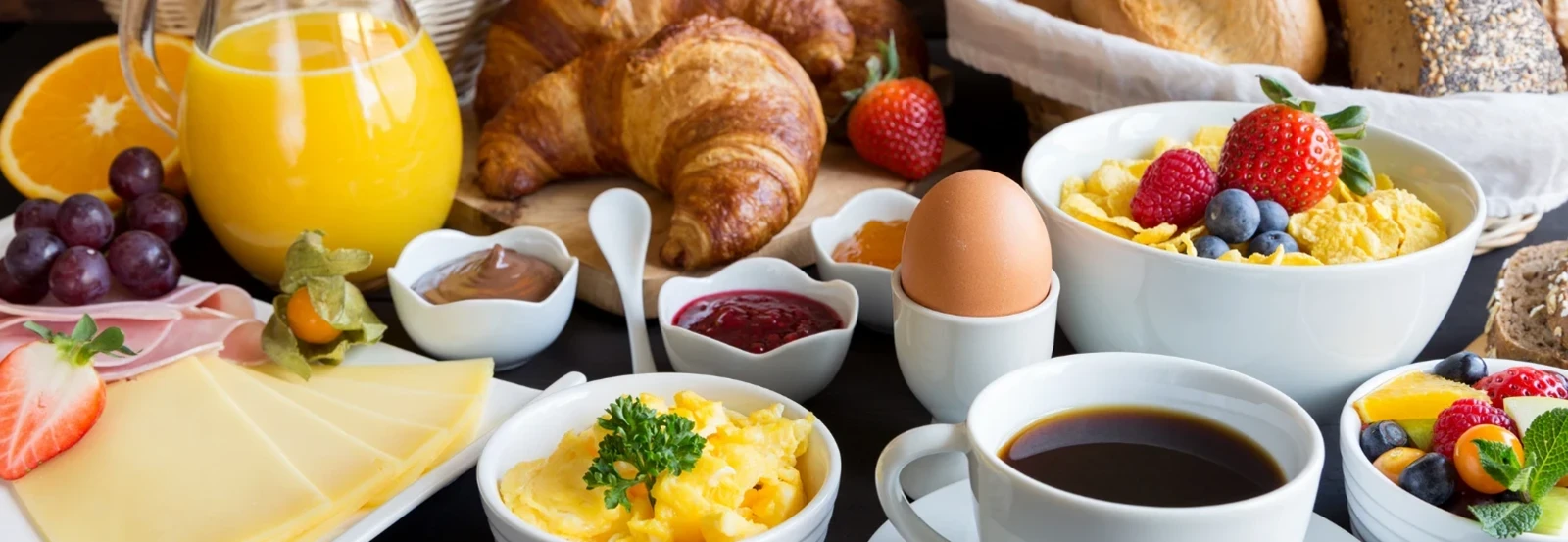 A breakfast spread with coffee, juice, bread, croissants, eggs, fruit, cheese, and cereal.