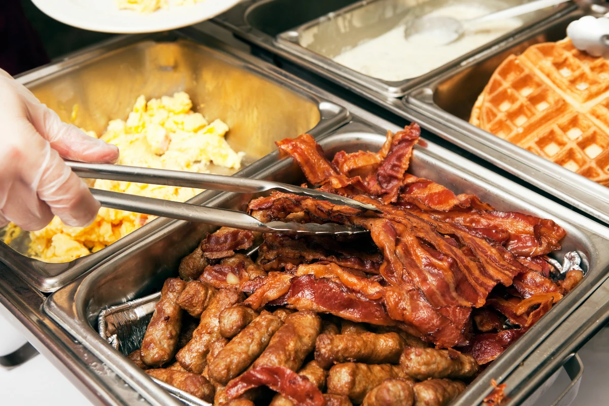 Buffet trays with bacon, sausage, scrambled eggs, and waffles, with tongs serving bacon.