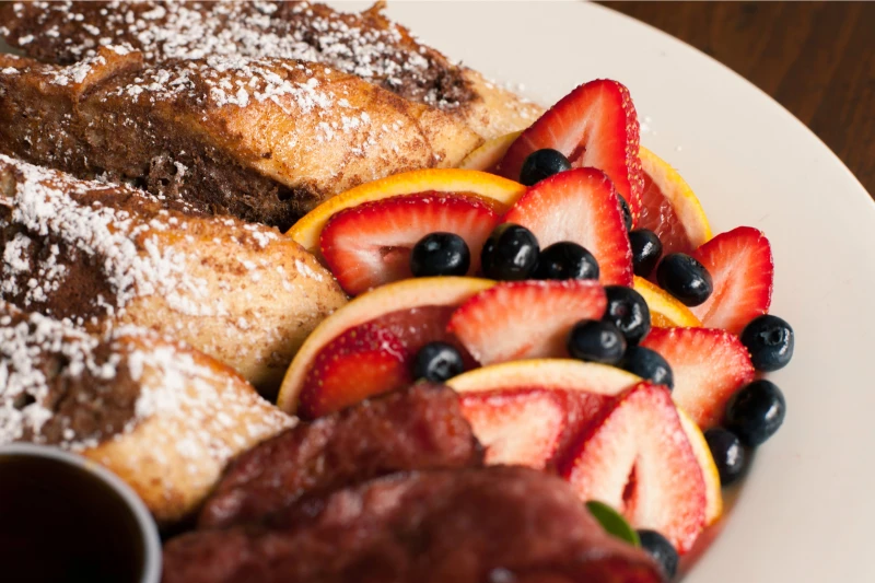 French toast with powdered sugar, bacon, orange slices, strawberries, and blueberries on a plate.