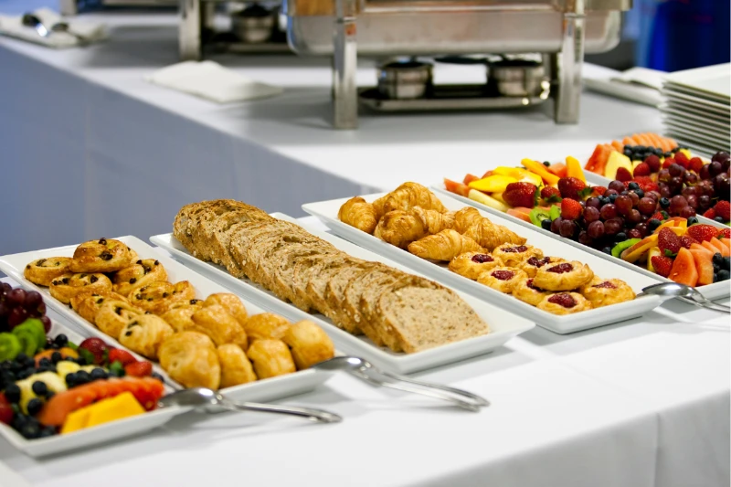 Platters of assorted pastries, breads, and fresh fruit on a white tablecloth at a buffet.