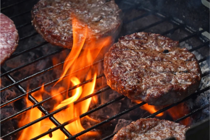 Beef burger patties cooking on a grill with flames rising between the metal grates.