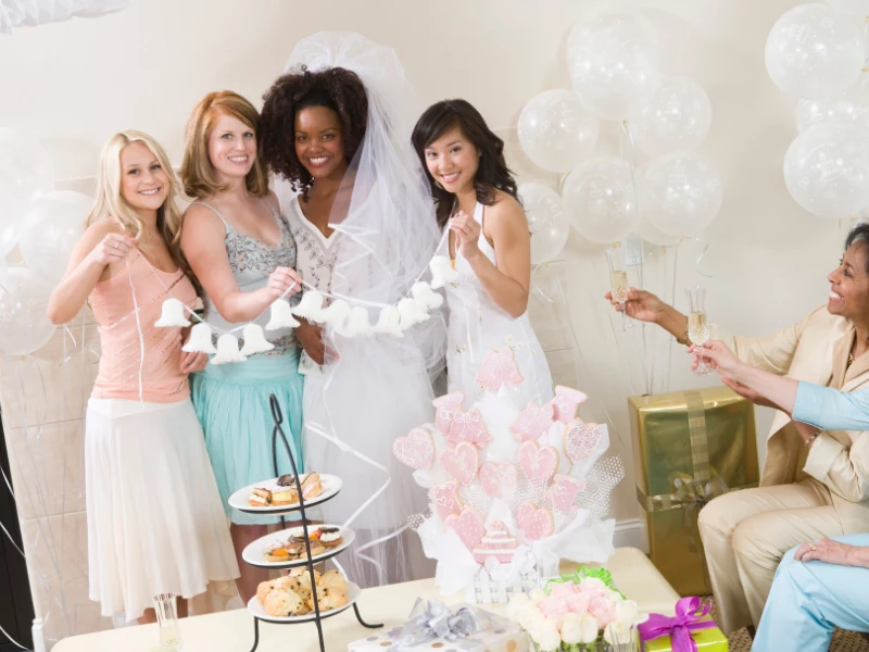 Four women in party dresses celebrate with decorations and treats, holding drinks and smiling.