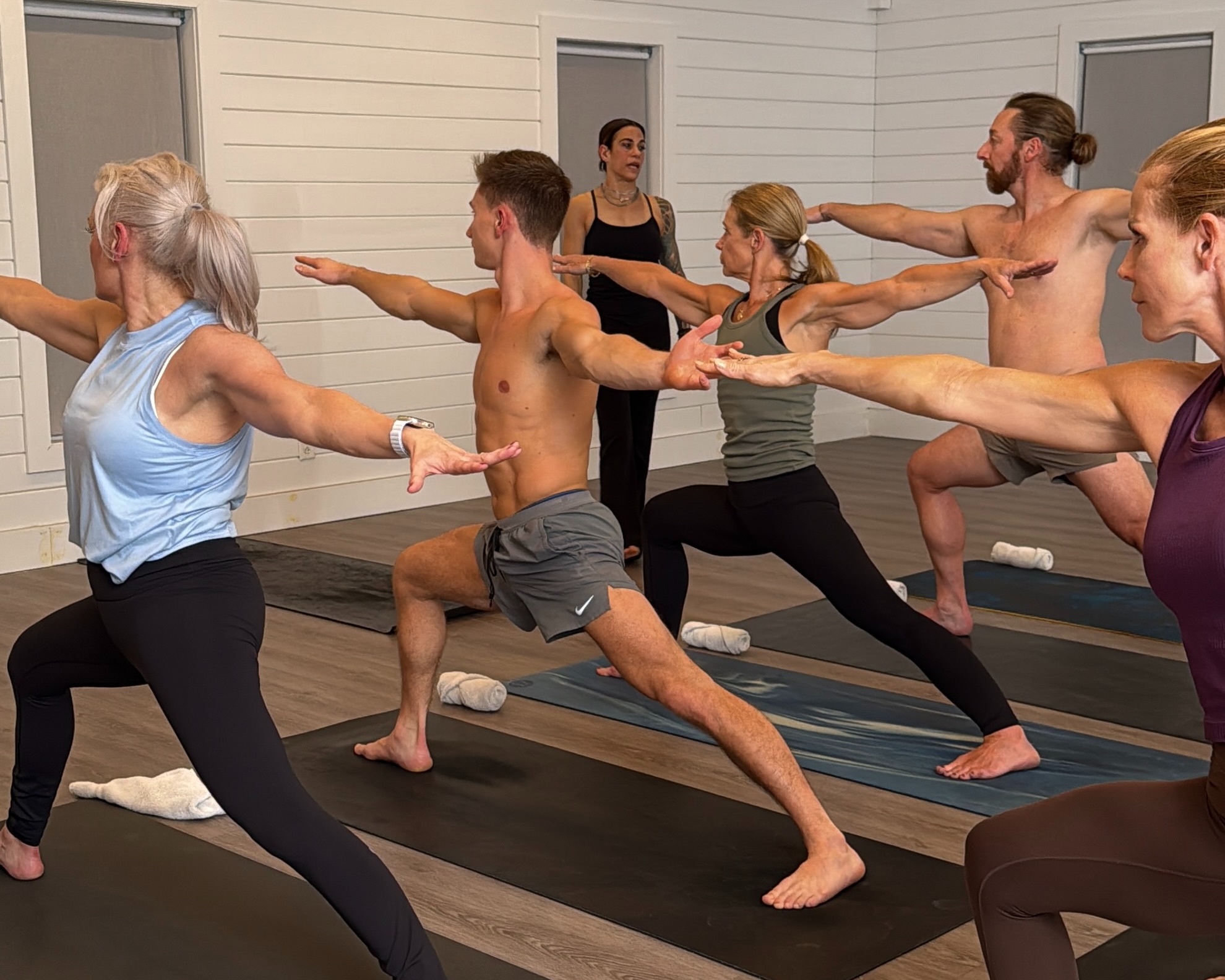 Five adults in a yoga class hold warrior poses on mats as an instructor guides them.