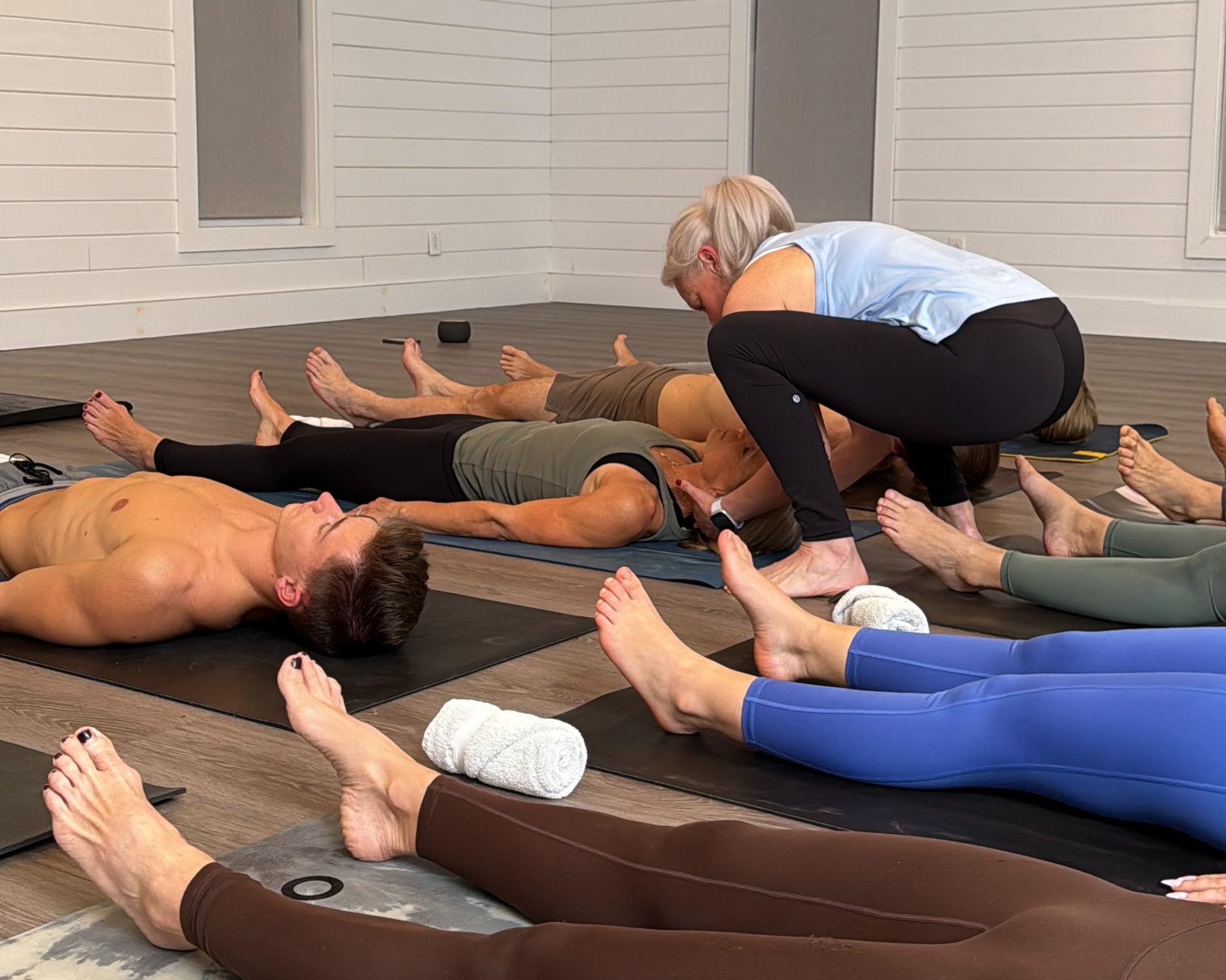 A yoga instructor assists a class of people lying on mats in a restful pose indoors.