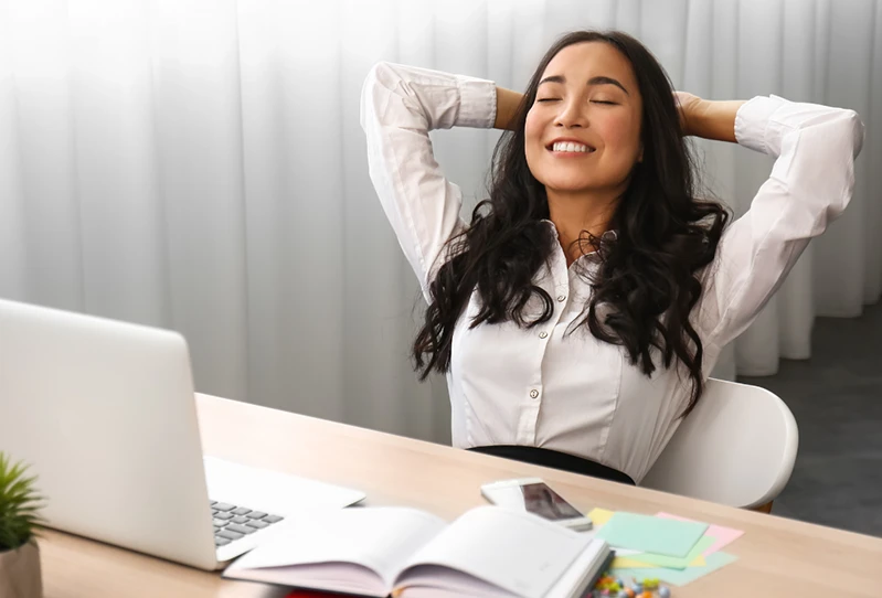 Smiling woman in business attire relaxes at her desk with hands behind her head.
