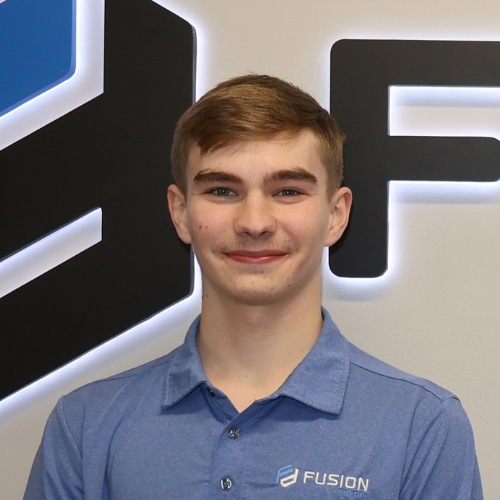 A bearded man in a blue blazer smiles in front of a logo on a wall.