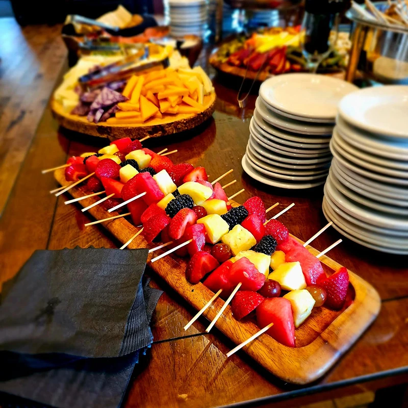 Wooden tray of fruit skewers on a table beside stacked plates, cheese, and napkins.