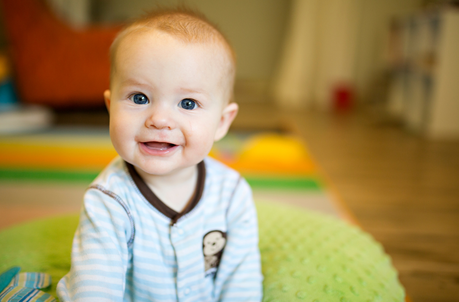Smiling baby in striped pajamas sits on a green cushion in a brightly lit room.