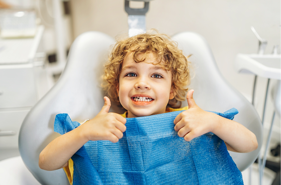 Smiling child in a dentist chair giving two thumbs up, wearing a blue dental bib.