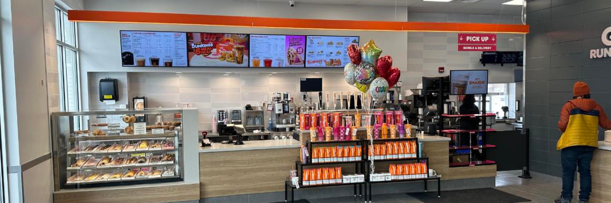 Dunkin’ Donuts counter with pastries, menu screens, balloons, and one customer waiting.